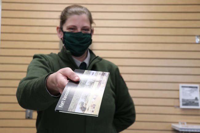 Smiling female park ranger with hair pulled back holds brochure in foreground with arm extended toward camera. In background is a yellow-beige wall with horizontal slats.