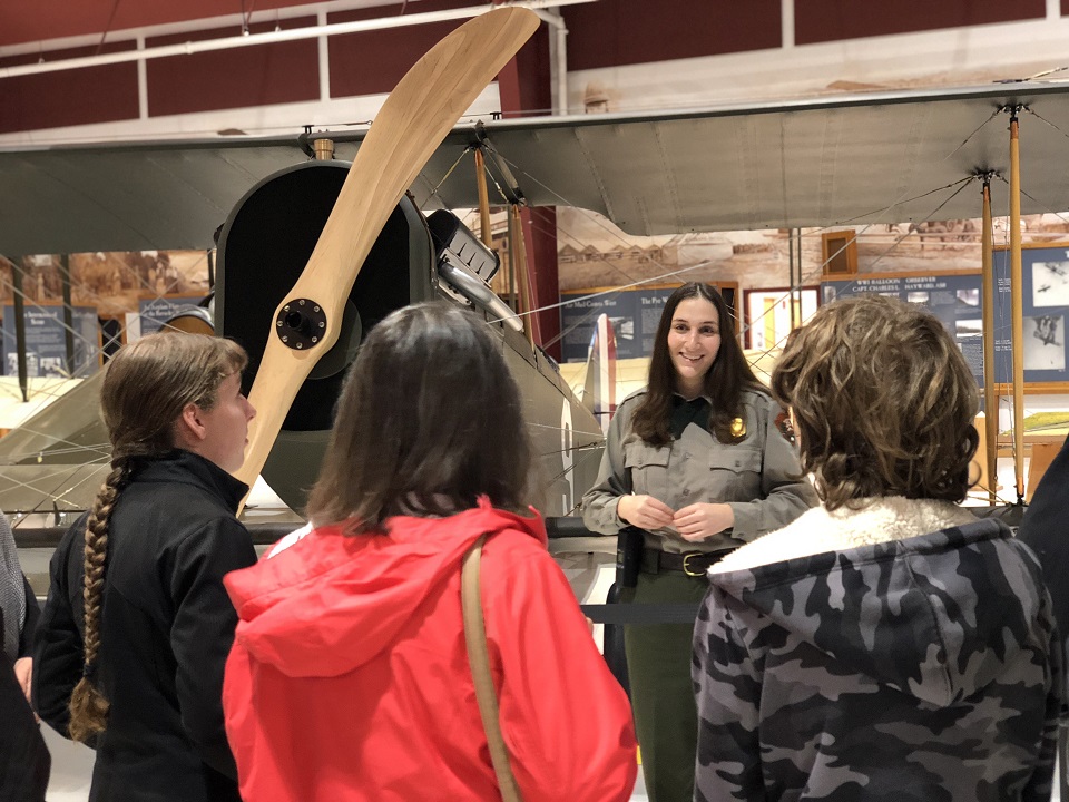 A woman in a park ranger uniform talks to people in front of a Curtiss Jenny aircraft.