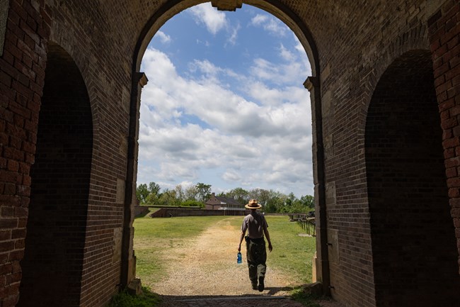A park ranger walks out of a shadowy archway into a sunlit parade ground.