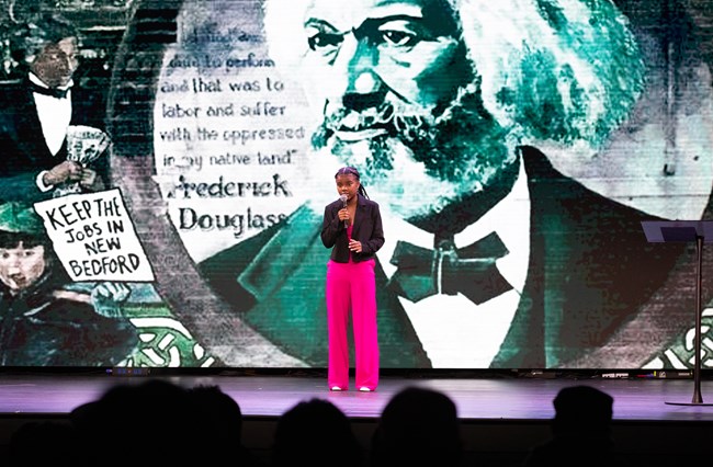 A young woman wearing a pink suit performs a speech on stage.
