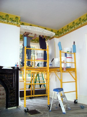 Two men stand on scaffolding and apply wallpaper to a room