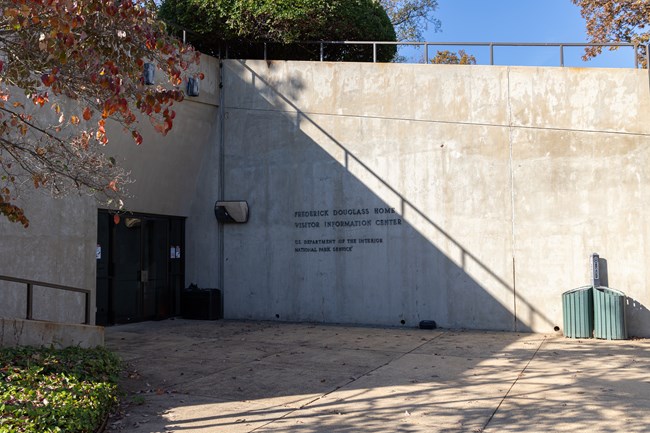 Two cement walls intersect to form the exterior of a visitor center. Double doors sit closed toward the left.