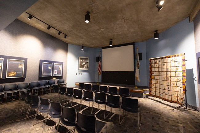Chairs lined up in three rows face a blank screen. A quilt hangs on the right and posters hang on the wall to the left.