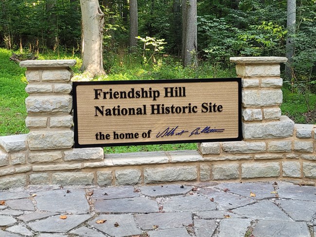 A sign reading Friendship Hill National Historic Site the home of Albert Gallatin. Trees and grass in background, slate in forground, sandstone columns bracketing sign