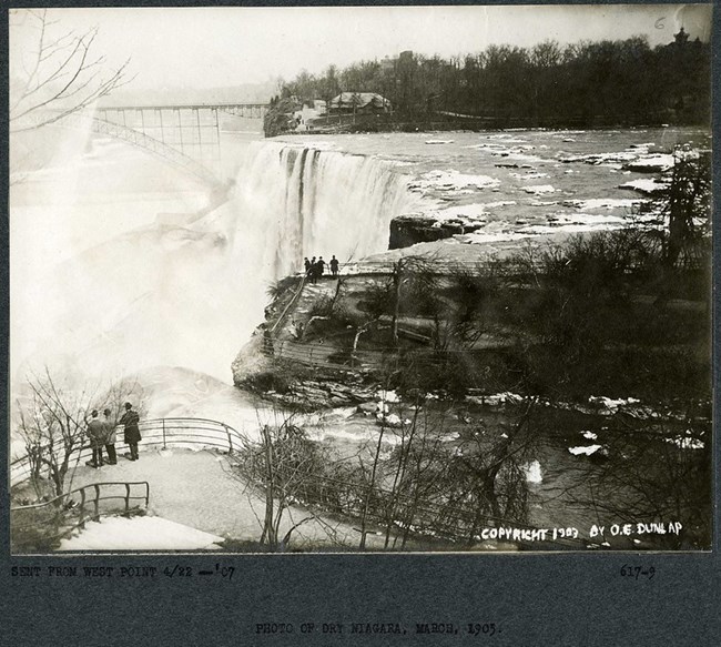 Black and white photograph of edge of waterfall. There are railings along the rock edges with few people standing near them. On the other side of the falls is a bridge, as well as a dense forest.