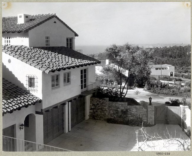 Black and white photograph of home in Spanish style with tile roof. In the distance are similar houses, many trees, and the ocean