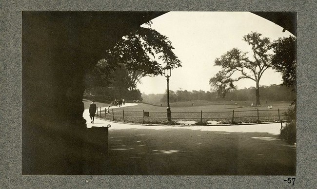 Black and white photograph of view from under an archway looking out onto a large open green space with walking paths surrounding it. Trees are planted along the edges, with only one in the central area.