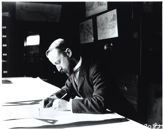 Black and white photograph of man in dark suit with light, tight hair hunched over a table, looking at a piece of paper, holding a pencil to it.