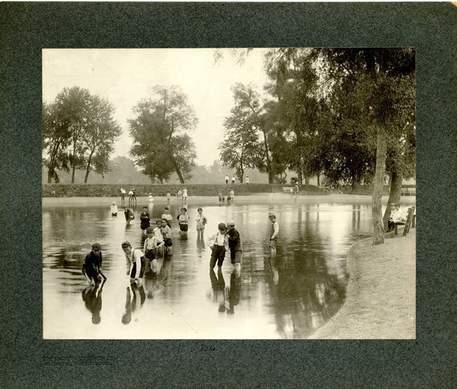 Black and white image of group of young children playing in body of water lined with brick wall and some trees along the wall.
