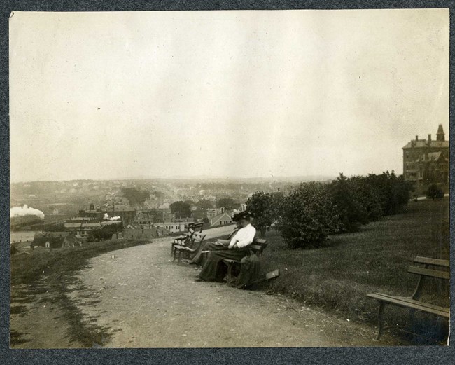 Black and white of top of hill with homes on the bottom, witha bench along a dirt path with a man sitting on the bench.