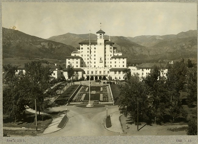 Black and white photograph of large white symmetrical building with a manicured garden out front and the peaks of mountains rising behind it.