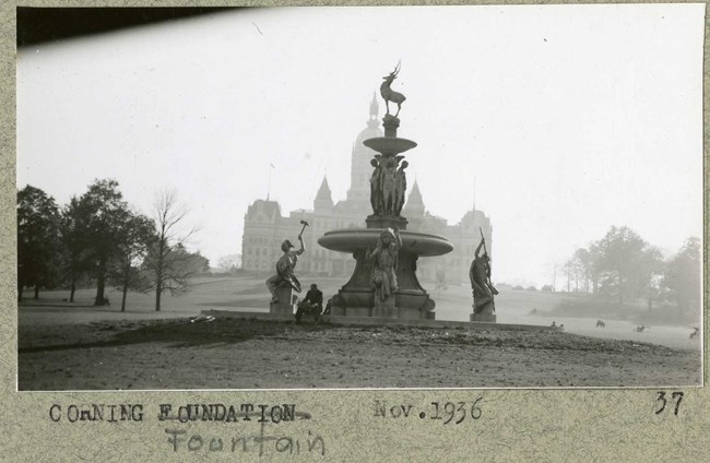Black and white of sculpture fountain with woman draped over it on large grassy area with trees on side and a building in the distance
