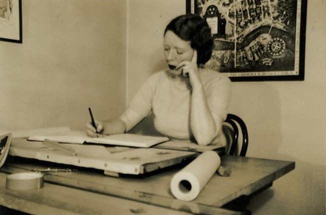 Black and white photograph of woman seated at desk hunched over papers with a pen in her hand.