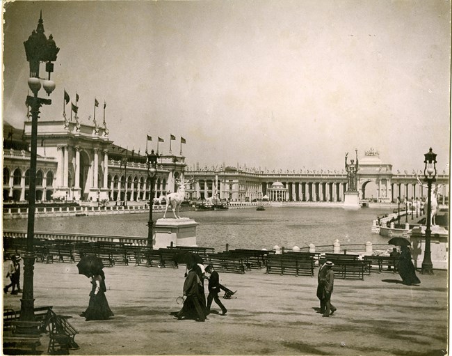 People walk along edge of water with palatial white large buildings rising around the water.