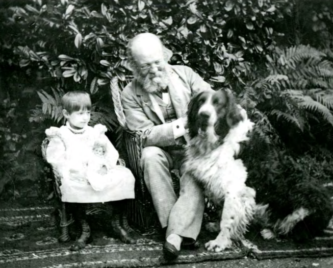 Black and white photograph of elderly man with thick white beard and bald head, though thick hair on the bottom, sits outside in front of bushes on top of a rug with a dog by his lap. A small child is also seated beside them.