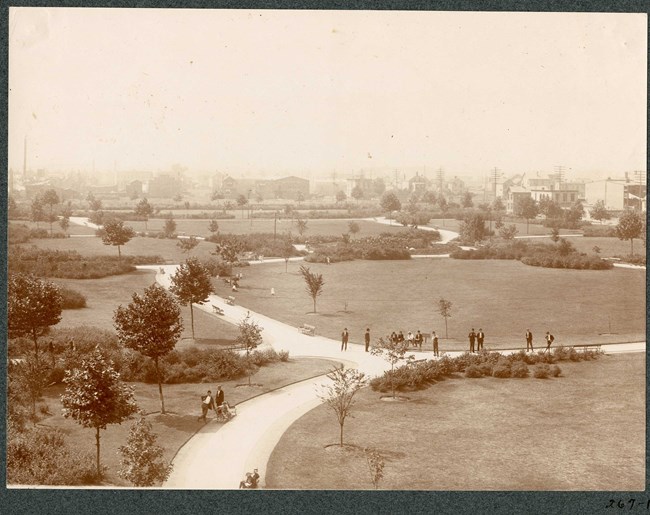 Black and white of aerial view of park with curving paths and some plantings along the edges, with some people on the path and buildings outside the park.