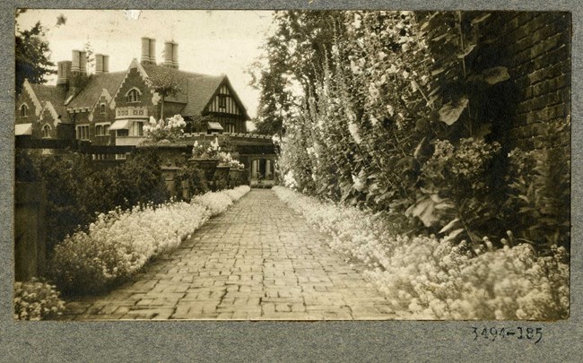 Black and white photograph of brick path leading to large home. The path is lined by flowers, and on one side is a wall with vines, the other with more plantings, some in vases.