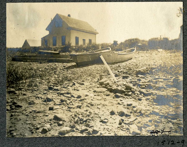 Black and white of rocky area with rowboats and a home on the rocks with trees in the distance.