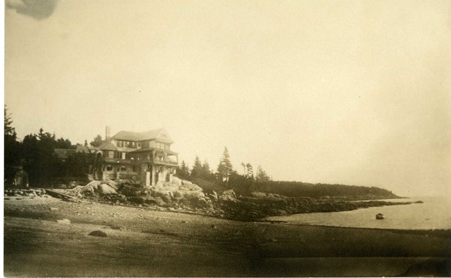 Black and white photograph of large home atop a pile of rocks on the shores of a beach. Behind the home are large trees, and in front is the sandy shore
