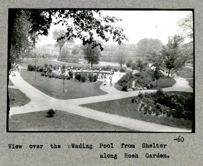 Black and white aerial view of paths cutting through grassy area with trees around the path edge and a small body of water with many people playing in it