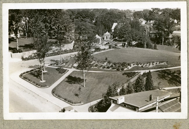 Black and white of rectangular flat park with paths cutting through, few trees spread out, some plants, and homes in the distance.