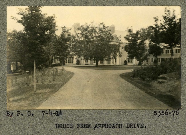 Black and white of driveway leading to large house with a fork, with open green circle with one tree at the center. Trees line the sides of the driveway.