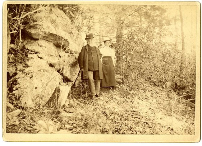 Black and white photograph of older man and young woman standing in the woods in front of a large rock.