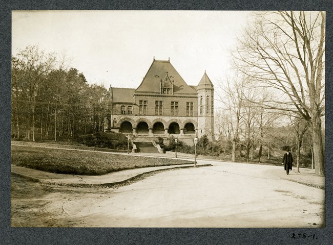 Black and white of dirt road leading to building on a hill with man walking along the road. The building has five arches, is made of stone, and has steps leading up.