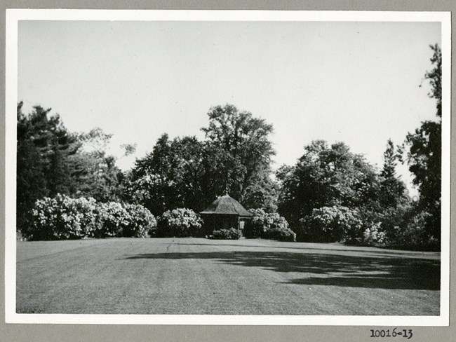 Black and white photograph of large grassy area with trees and shrubs on the edges, with a small structure at the end.