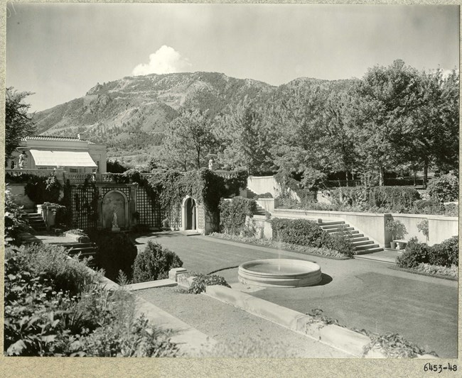 Black and white photograph of garden with walls covered in vines and arches for statues. The center of the garden is a rectangular grassy area, with a circular fountain.
