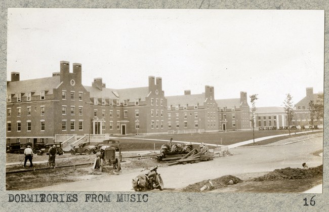 Black and white of construction on school, with road, materials left on road, grassy area with people and cars, and a row of brick buildings which all look the same. In front of the buildings is a grassy area.