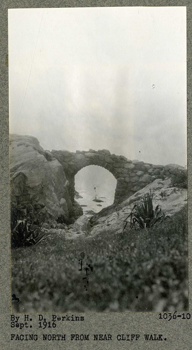 Black and white photograph of stone arch going between two large rocks leading towards the ocean