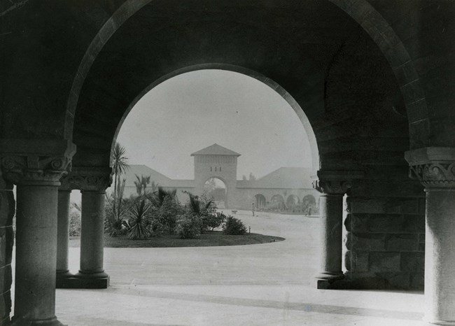 Black and white looking through arch to open area with circle of grass, walking paths, and another building in the distance