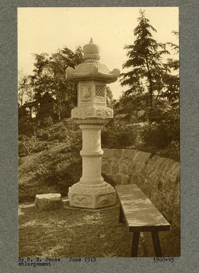 Black and white photograph of large white ornamental statue in garden with rock wall and plantings behind it, and a wooden bench next to it.