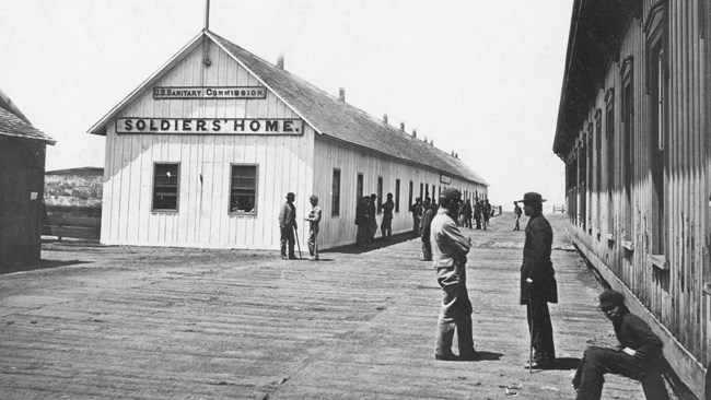 Black and white photograph of large wooden building very basic in design, with a United States Sanitary Commission Soldiers Home on one side. Many men stand outside the building.