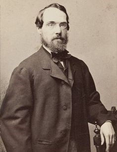 Black and white photograph of man with dark head of hair and a thick beard with oval glasses wearing a suit and posing with a chair