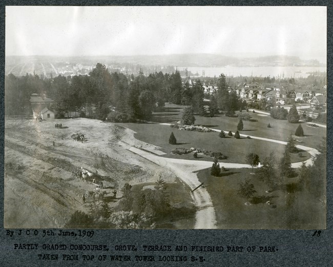 Black and white aerial of park under construction with looping path around open green space with homes and a dense stand of trees in the distance. One half of the park is all dirt