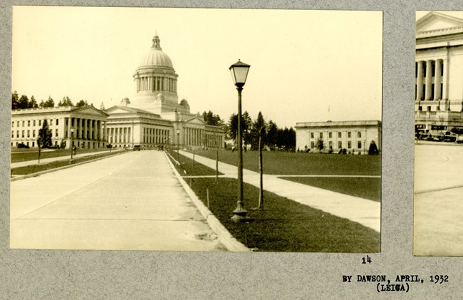 Black and white of concrete path with grass on both sides and walking path in grass, leading to a large white building with many columns and a dome.