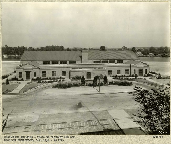 Black and white of symmetrical building with curving drive in front with grassy area and plantings, with a railroad track in front and water behind.