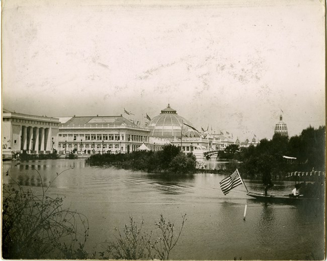 Black and white of body of water with boat on it with USA flag and large buildings in background.