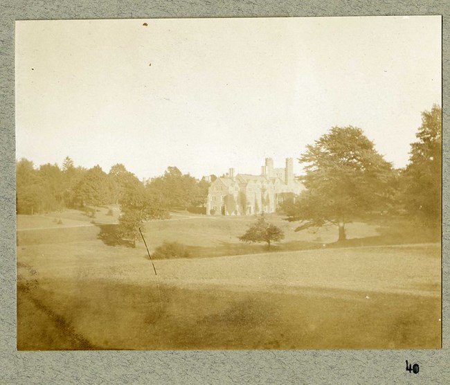 Black and white photograph of castle-like building in a large field with trees surrounding the edges, with some in the open space