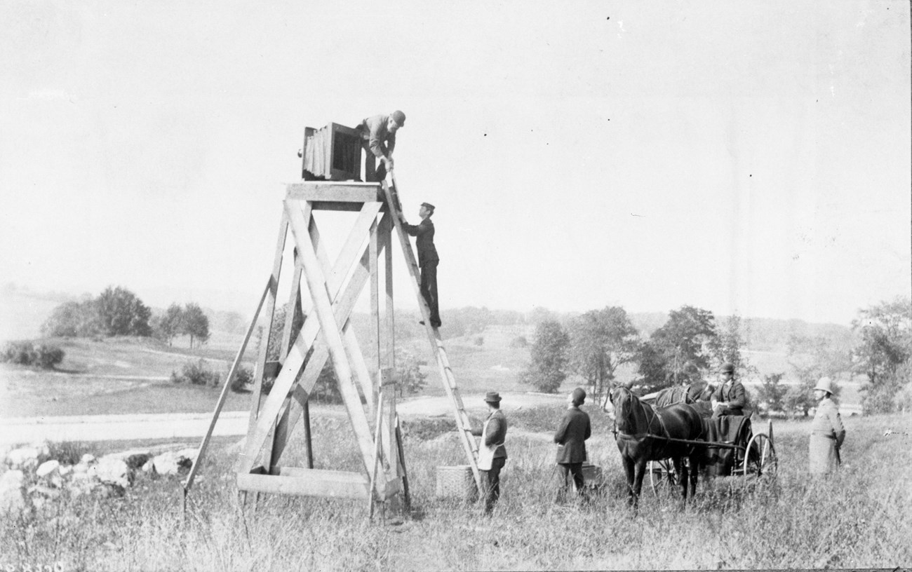 Black and white of large wooden tower with camera on top, people climbing up, horse and people at bottom.