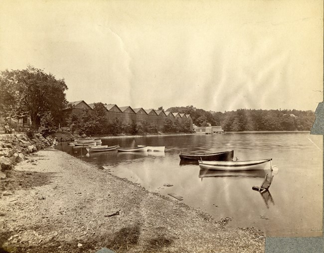 Black and white of pond shore with boats and a building in distance