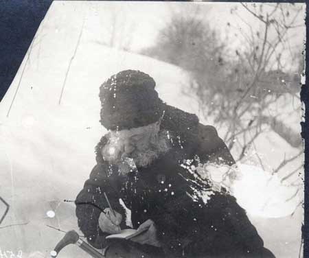 Black and white image of Olmsted in winter hat and jacket sitting outside in the snow while writing in a journal.