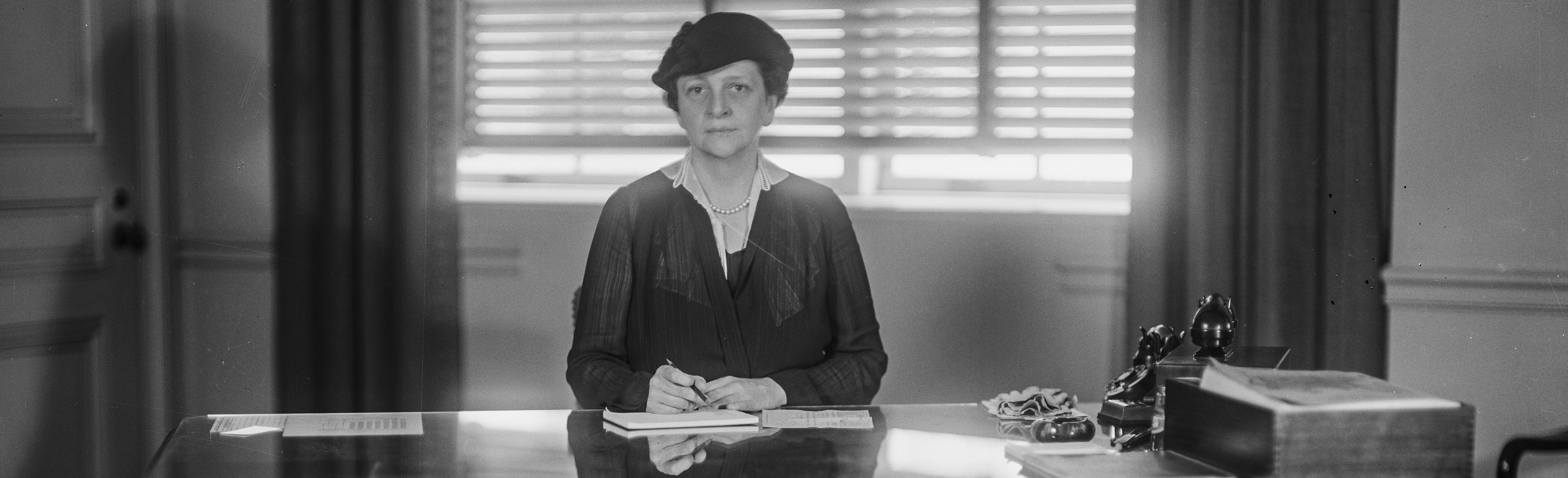 Frances Perkins, seated directly at the center of the frame behind a desk, looking directly at the camear, with a pen in hand as if about to sign the document in front of her.