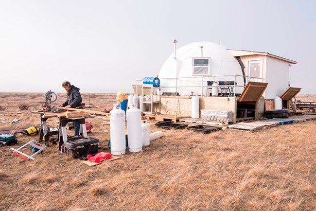 A man operating a table saw in front of a small structure on the coastal plain