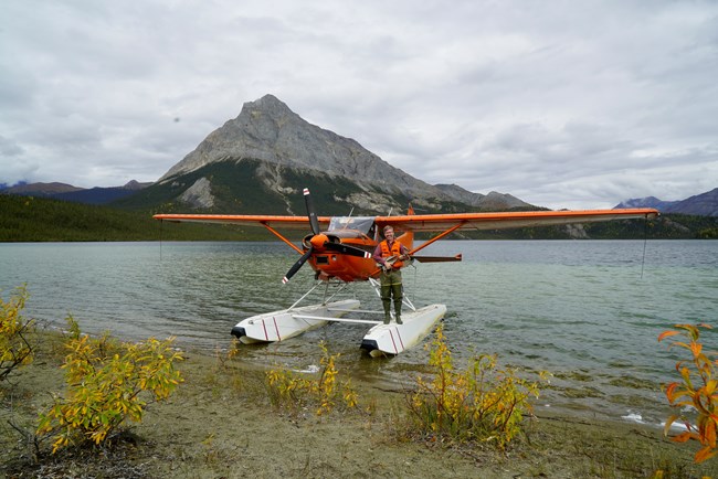 a pilot stands, holding a small anchor, on his bush airplane equipped with floats. The plane is on a dark blue lake with tall mountains in the background and cloudy skies.