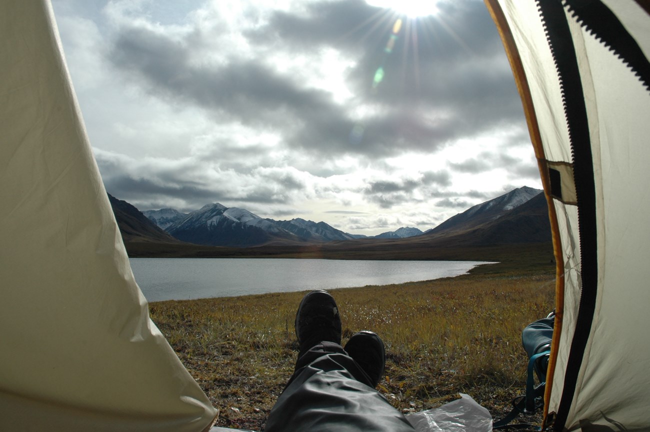 Feet sticking out of an open tent with a view of a large blue lake and tall mountains behind