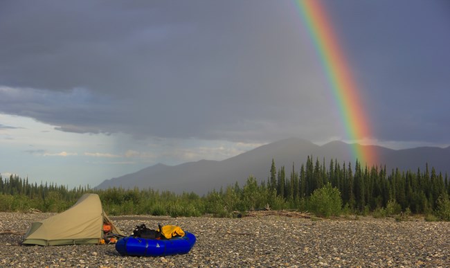 Green and Tan Tent with man's head peaking out of the flap, next to a blue packraft, situated on the stony riverbank. A rainbow appears in the sky in front of an incoming storm against mountains the background