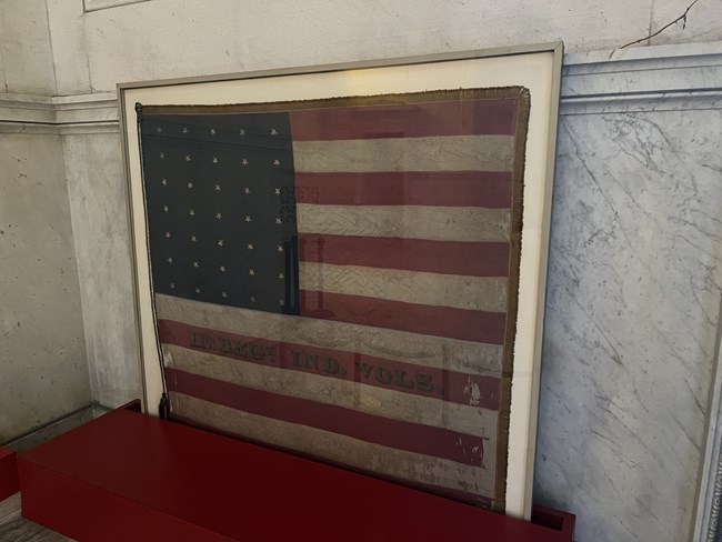 An old flag on display in a case against a marble wall.